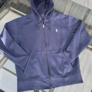 Marinblå hoodie från Polo Ralph Lauren - Säljer en marinblå hoodie från Polo Ralph Lauren med dragkedja och huva. Tröjan har fickor framtill och den klassiska vita loggan broderad på bröstet. Perfekt för en avslappnad stil. Ser urtvättad ut på bilderna p.g.a ljuset men i verkligheten är den i mycket bra skick☺️ Det ärevarvs fråga på😊