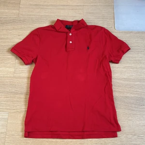 Röd pikétröja från Polo Ralph Lauren - Säljer en klassisk röd pikétröja från Polo Ralph Lauren i slim fit-modell. Den är köpt på nk för ungefär ett år sedan men har knappt använt den för den inte passat, har tyvärr inte kvittot kvar. Tröjan har korta ärmar, krage och två knappar framtill samt den ikoniska broderade loggan på bröstet. Perfekt för en stilren och sportig look. 
