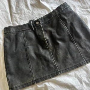 Svart skinnkjol från PrettyLittleThing - Cool svart/khaki skinnkjol från PrettyLittleThing med en kort passform. Kjolen har en knapp och dragkedja framtill och tydliga sömmar som ger en snygg look. Perfekt att styla med en enkel topp. 