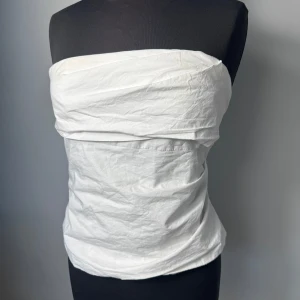 Vit tubtopp med dragkedja - Säljer en stilren vit tubtopp med dragkedja bak. Toppen är axelbandslös och har en enkel, clean design som passar perfekt till sommaren. Materialet känns lätt och bekvämt mot huden. Storlek M och är använd bara fåtal gånger💕😊