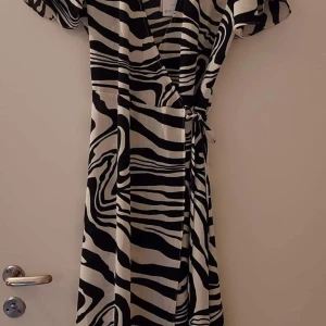 Zebramönstrad omlottklänning - Ny omlottklänning med zebramönster i svart och vitt med lappen kvar. Klänningen har korta ärmar och knyt i midjan för en smickrande passform. Perfekt för dig som vill sticka ut med ett trendigt djurmönster. 