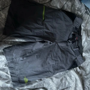 Grå shorts från The North Face - Mörkgrå shorts från The North Face med diskret limegrön logga på benet och praktiska fickor. Modellen har bälteshällor och är tillverkad i ett lätt och slitstarkt material, perfekt för utomhusaktiviteter.