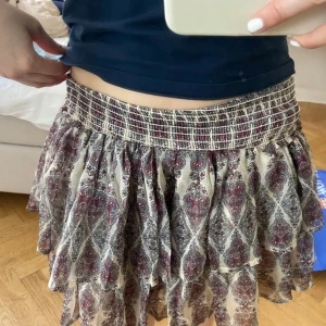 Mönstrad volangkjol med smock - Säljer en kort volangkjol med bred smock i midjan. Kjolen har ett bohemiskt mönster i vinrött, grått och beige och är luftig med flera lager volanger. Perfekt för dig som gillar en avslappnad och trendig stil.