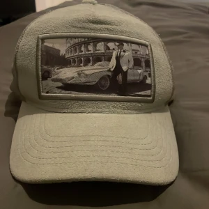 Natone grish keps - Snygg keps med fototryck framtill som visar en klassisk bil och person framför Colosseum. Kepsen har mesh på sidorna och broderad Natone-logga på sidan. Perfekt accessoar för bilentusiasten.