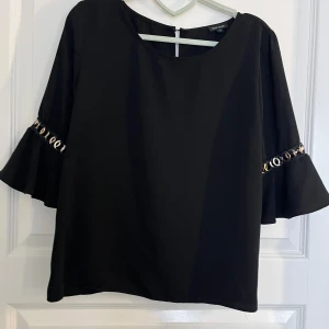 Svart blus med volangärm från River Island - Svart blus från River Island med vida volangärmar och dekorativa guldfärgade ringdetaljer vid ärmslutet. Blusen har rund halsringning och är tillverkad i ett mjukt tyg som faller snyggt.