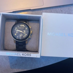 Svart och guldig klocka från Michael Kors - Säljer en snygg Michael Kors klocka, modell MK5191. Klockan har svart armband och urtavla med guldiga detaljer och visare. Flera små urtavlor och robust design. Perfekt accessoar för att lyfta din stil.