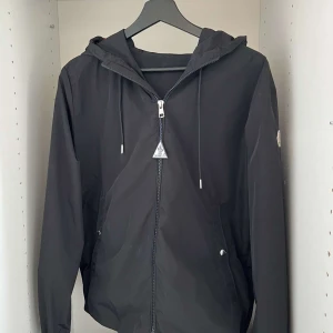Moncler vindjacka  - Säljer en svart Moncler windbreaker med huva och dragkedja framtill. Jackan är köpt på NK och är helt äkta doch finns inget kvitto då det försvann under en flytt. Perfekt för blåsiga dagar och enkel att matcha med det mesta. Säljer nu för att jag inte använder den längre men den är i väldigt bra skick