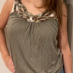 Olivgrön linnetopp med dekorativt halsband - Säljer en olivgrön linnetopp med unikt, dekorativt halsband i metallicfärgade detaljer vid ringningen. Toppen är ärmlös och har en avslappnad passform, perfekt för varma dagar.