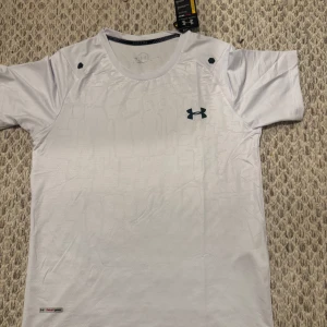 3st Under armour - Snygg t-shirt från Under Armour med diskret mönster och logga på bröstet. Finns i vitt, svart och grått. Perfekt för träning eller sportiga dagar. Tillverkad i ett lätt och snabbtorkande syntetmaterial. Klassisk passform och korta ärmar.