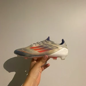 Adidas fotbollsskor med röda och blå detaljer - Säljer ett par Adidas fotbollsskor i vitt med röda och blå detaljer. Skorna har snörning och är designade för spel på gräs med dobbar undertill. Lätta och smidiga med en sportig look, perfekta för dig som vill ha snabbhet på planen. Knappt använda 