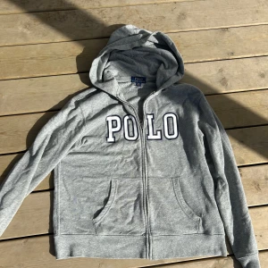 Grå hoodie från Polo Ralph Lauren - Säljer en grå hoodie från Polo Ralph Lauren med dragkedja och stor POLO-text framtill. Tröjan har huva och två fickor på magen. Perfekt för en avslappnad stil. Pris kan diskuteras. 