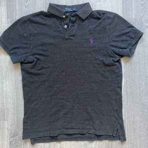 Svart piké t-shirt från Polo Ralph Lauren - Klassisk svart piké t-shirt från Polo Ralph Lauren med lila broderad logga på bröstet. Slim fit-modell med korta ärmar och krage med två knappar. Perfekt för en stilren och avslappnad look.