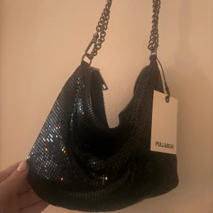 Svart glittrig axelväska från Pull&Bear - Säljer en svart glittrig axelväska från Pull&Bear med kedjehandtag och dragkedja. Perfekt för att lyfta din outfit med lite extra bling. Väskan är liten och smidig, passar när du vill bära det viktigaste köpt för 330.