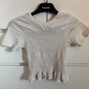 Vit croppad topp med volangkant - Säljer en söt vit croppad topp från Gina young med smockat parti framtill och volangkant nedtill. Toppen har korta ärmar och rund halsringning. Perfekt till sommaren!