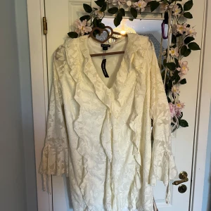 Ivoryfärgad volangklänning från Boohoo - Säljer en ljus, ivoryfärgad klänning från Boohoo med blommigt jacquardmönster och volangdetaljer längs v-ringningen och ärmsluten. Klänningen har lång ärm och en romantisk känsla, perfekt för speciella tillfällen. Den är normal i storleken förutom längden som är ganska kort.