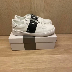 Valentino Garavani vita sneakers med svarta detaljer - Säljer ett par vita sneakers från Valentino Garavani med svart band och nitar på sidorna. Skorna har snörning och en platt sula. Perfekta för dig som gillar stilrena men ändå unika detaljer.