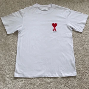 Vit t-shirt från Ami Paris - Stilren och klassisk vit t-shirt från franska AMI Paris, med det ikoniska röda hjärtat på bröstet. Tillverkad i 100% mjuk bomull, perfekt att bära både till vardags och vid mer uppklädda tillfällen. T-shirten är aldrig använd och pris kan diskuteras. Skriv om ni har frågor 