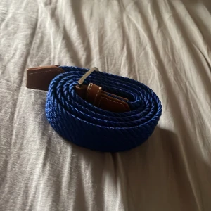 Blått flätat bälte med läderdetaljer - Säljer ett blått flätat bälte med bruna läderdetaljer och silverfärgat spänne. Bältet har en snygg och modern look som passar till jeans eller chinos. Perfekt accessoar för att lyfta din outfit.