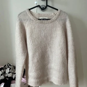 Beige stickad tröja - Mysig beige stickad tröja med rund halsringning och långa ärmar. Tröjan har en enkel och klassisk design med ribbade muddar vid ärmslut och nederkant. Perfekt för lager-på-lager under kyliga dagar.