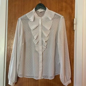 Volang blus - Vit, långärmad blus med volang på bröstet. På bilden ser den gråare ut i verkligheten eftersom att den är genomskinlig men fotas mot en mörk dörr! Storlek M. 🫶🏻