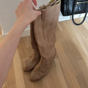 Beiga höga stövlar med klack - Säljer ett par snygga beiga stövlar i skinn med klack och rund tå. Stövlarna är knähöga och har en stilren design utan synliga detaljer som snörning eller dragkedja. Perfekta för att lyfta din outfit! Bootsen har lite vattenskador men annars i bra skick🥰