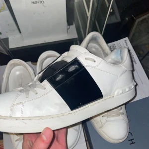 Valentino Open Sneakers - Säljer ett par snygga och stilrena Valentino Garavani Open sneakers i storlek 44. Klassisk modell med svart band i läder över vristen – en riktig designikon som lyfter vilken outfit som helst. Skorna är använda men i gott skick. Visst bruksslitage finns (se bilder), bland annat på sulorna och lite veck i lädret – men inga skador eller defekter. Allt är rengjort och redo för ny ägare!  Allt og utom kvitto medföljer!                 📦 Skickas spårbart – hör av dig vid frågor! 