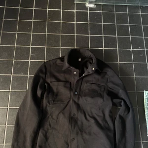 Svart overshirt med knappar - Säljer en svart overshirt med klassisk krage och två bröstfickor. Jackan har knäppning framtill och är långärmad. Perfekt för lager-på-lager och enkel att styla till olika outfits.