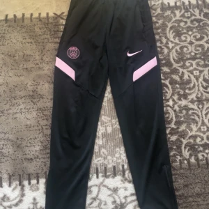 Svarta träningsbyxor från Nike x PSG - Svarta träningsbyxor från Nike i samarbete med Paris Saint-Germain. Byxorna har rosa detaljer, PSG-logga och Nike Swoosh på framsidan. Tillverkade i syntetmaterial med smal passform och dragkedjor vid bensluten.