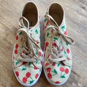 Beige sneakers med körsbärsmönster från ASOS - Säljer ett par beige sneakers från ASOS med färgglatt körsbärsmönster i rött och grönt. Skorna har vita sulor och beige snörning. 