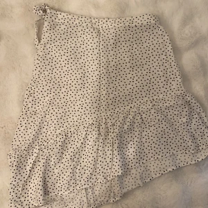 Vit prickig kjol från Kappahl - Supersöt vit kjol från Kappahl med svarta prickar och volang nertill. Kjolen har omlottknytning i midjan och är perfekt för en somrig look. Lätt och luftig modell som passar till många olika stilar.
