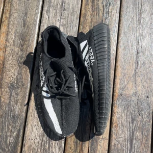 Svarta Yeezy-inspirerade sneakers - Svarta sneakers med vit rand och textdetaljer på sidan, inspirerade av Yeezy Boost 350. Skorna har snörning, stickad ovandel och en tjock, räfflad sula. Perfekta för dig som gillar streetstyle och vill sticka ut med en unik look.