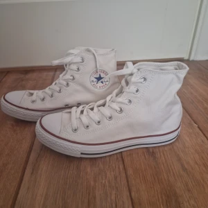 Vita Converse All Star high sneakers - Klassiska vita Converse All Star high sneakers med rund tå, snörning och röd rand längs sulan. Skorna är i canvas med gummisula och har den ikoniska loggan på sidan. Perfekta för en avslappnad och cool stil. 