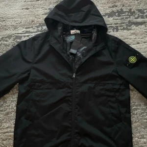 Stone Island Svart vindjacka - Säljer en svart vindjacka från Stone Island med huva och dragkedja framtill. Passar bra i sommarkvällar.