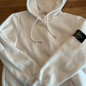 Vit hoodie från Stone Island - Vit hoodie från Stone Island med klassisk logopatch på vänster ärm. Tröjan har huva med snörning och ribbade muddar vid ärmslut och nederkant. Mjuk och skön, perfekt för en chill look.