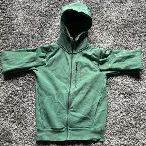 Grön hoodie från Sail Racing XS - Grön hoodie från Sail Racing i storlek XS. Tröjan har hel dragkedja framtill, huva med snörning och en bröstficka med dragkedja. Tillverkad i mjukt material med fleece på insidan, perfekt för kyliga dagar.