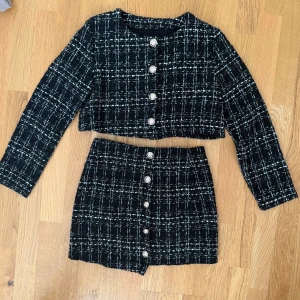 Svartvit rutig tweed kostymset - Snyggt tvådelat kostymset i svart och vitt tweedtyg med rutigt mönster. Kavajen är croppad med långa ärmar och dekorativa silvriga knappar framtill. Kjolen är kort och har matchande knappar längs hela framsidan. Perfekt för en trendig look!  OBS: Själva ziptaggen på kjolens dragkedja är trasig. Jag använder hårnålar för att dra upp den. Dragkedjan fungerar, men köparen får hitta en egen lösning om man vill byta ut eller laga den.