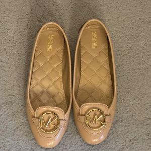 Beige ballerinaskor Michael Kors - Snygga beige ballerinaskor från Michael Kors i skinn med quiltad innersula och rund tå. Framtill sitter en guldig MK-logga som ger en lyxig touch. Perfekta för dig som gillar stilrena och klassiska detaljer. Bara använt en gång🩷🩷