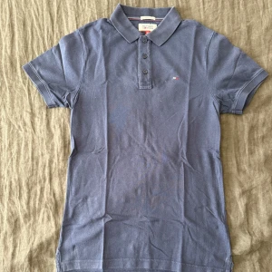 Marinblå pikétröja Tommy Hilfiger - Snygg mörkblå pikétröja från Tommy Hilfiger Denim med klassisk krage och tre knappar framtill. Tröjan har korta ärmar och en liten broderad logga på bröstet. Perfekt för en clean och stilren look.