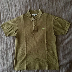 Mossgrön pikétröja från Lacoste - Snygg mossgrön pikétröja från Lacoste i klassisk passform. Tröjan har korta ärmar, krage med två knappar och den ikoniska krokodil-loggan broderad på bröstet. Tillverkad i bomull som känns skön mot huden och passar perfekt till jeans eller shorts.