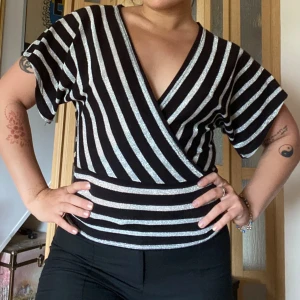 Glittrig omlottopp från Vila - Säljer en svart och silverglittrig omlottopp från Vila med v-ringning och korta ärmar. Toppen har ett randigt mönster med både lodräta och vågräta ränder och en croppad passform. Perfekt för dig som vill sticka ut lite extra!