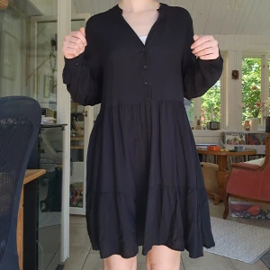 Svart volangklänning från Amisu - Svart kortklänning från Amisu med v-ringning, knappar framtill och långa ärmar. Klänningen har volanger i kjolen och en lös, flowy passform som ger en chill vibe. Perfekt för dig som gillar enkel men snygg stil.