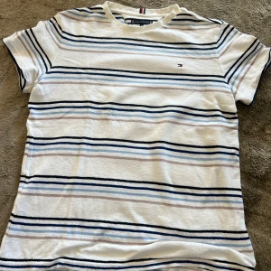 Randig t-shirt från Tommy Hilfiger - Snygg t-shirt från Tommy Hilfiger i vitt med tunna blå, svarta och beige ränder. Klassisk passform med korta ärmar och rund hals. Liten broderad logga på bröstet. Perfekt för en chill och stilren look.