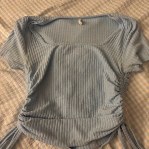 Ljusblå ribbad topp med knyt - Superfin ljusblå ribbad topp med kort ärm och fyrkantig ringning. Snygga knytband i sidorna som ger en trendig look. Perfekt att styla med jeans eller kjol för en chill vibe.