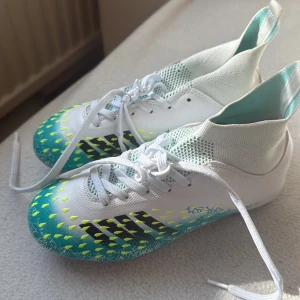 Adidas vita och turkosa fotbollsskor - Snygga fotbollsskor från Adidas i vitt och turkost med svarta ränder och gula detaljer. Skorna har snörning, stickad strumpliknande krage och mönstrad ovandel. Perfekta för dig som vill sticka ut på planen!