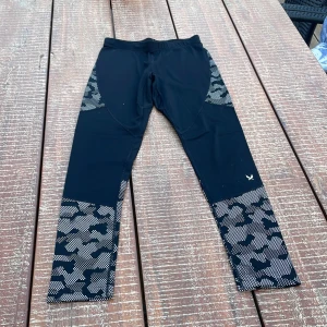 Svarta träningsleggings från SOC - Svarta leggings från SOC med grått camomönster på sidorna och nedtill på benen. Tillverkade i stretchigt syntetmaterial som andas, perfekta för träning. Tight passform och hög midja för en sportig look.