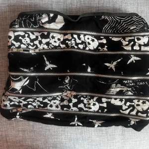 Svart miniskirt med dödskallemönster och dragkedjor - Cool svart kortkjol med vita dödskallar och benknotor som mönster. Kjolen har flera silvriga dragkedjor som detaljer och ger en edgy vibe. Perfekt för dig som gillar alternativ stil och vill sticka ut.