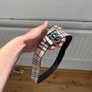 Burberry bälte - Snyggt Burberry-bälte med klassiskt rutigt mönster i beige, svart och rött på ena sidan och svart/grått på andra. Stort metallspänne med B-logga framtill. Bältet är gjort i skinn och har en bred, trendig form som sticker ut i outfiten.