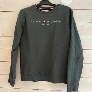 Mörkgrå/grön  sweatshirt från Tommy Hilfiger - Mörkgrå/grön sweatshirt från Tommy Hilfiger med klassisk rund hals och tryckt logga framtill. Tröjan har ribbade muddar vid ärmslut och nederkant samt en enkel, stilren design. Perfekt för en avslappnad och snygg look.