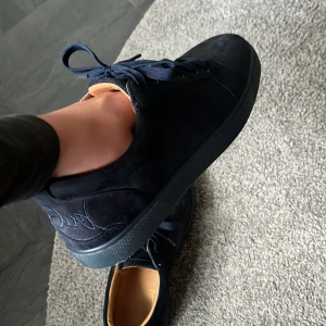 Mörkblå sneakers från Louboutin - Snygga mörkblå sneakers från Christian Louboutin i mocka med klassisk röd sula och broderad logga på sidan. Skorna har rund tå, snörning och en stilren design som sticker ut med den ikoniska röda undersidan. Saknar box.