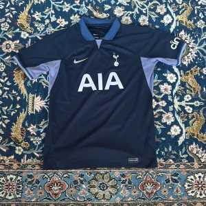 Tottenham Nike fotbollströja  - Snygg Tottenham Hotspur fotbollströja från Nike i Dri-FIT-material. Tröjan är marinblå med ljusblå detaljer på sidorna och kragen. Stor AIA-logga på bröstet och klubbmärke på vänster sida. Perfekt för dig som vill visa ditt stöd för Spurs. Tröjan har ingen skador eller liknande då den är väl omhändertagen. Tveka inte på att fråga om ni undrar nått!